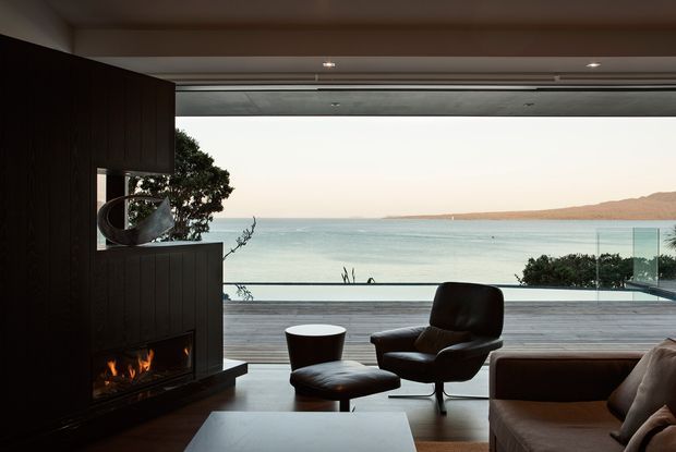 A sophisticated contemporary living room opening onto a broad timber deck with uninterrupted ocean views. The room features dark timber wall panelling and built-in cabinetry, a linear fireplace, leather lounge chairs with ottoman, a neutral sofa, a low coffee table, and expansive retractable glazing. The setting feels calm, refined, and strongly connected to the coastal landscape.