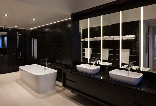 A spacious contemporary bathroom featuring a freestanding white bathtub, double white vessel basins on a long dark vanity, large illuminated mirrors, black mosaic wall tiles, light floor tiles, and recessed ceiling lighting. White towels are displayed on open shelving behind the vanity.