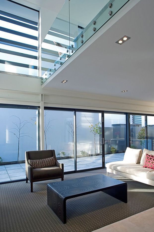Bright contemporary living room with double-height glazing, a glass-balustraded upper-level landing, recessed ceiling lights, and direct access to a paved courtyard. The room contains a dark upholstered armchair, white sofa, patterned cushions, and a low sculptural coffee table, with strong daylight entering through large sliding glass doors.