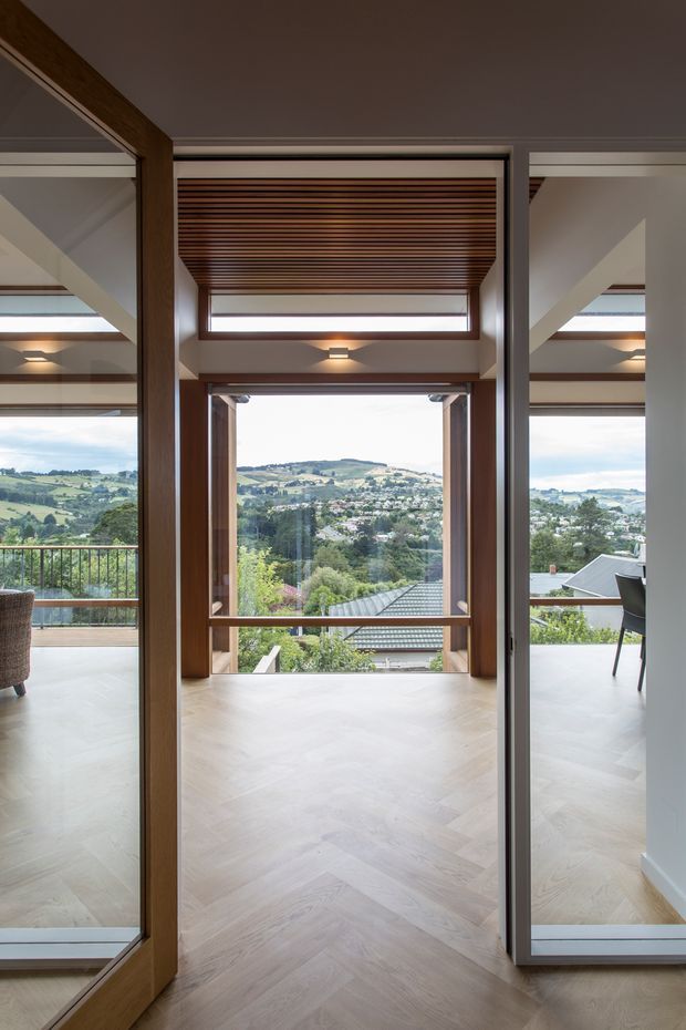 A refined contemporary interior viewed through a timber-framed opening toward a bright glazed living space and elevated landscaped suburban view. The room features pale herringbone timber flooring, extensive floor-to-ceiling glazing, warm timber structural framing, white walls, and a timber slatted ceiling detail. A balcony railing, outdoor greenery, and neighbouring houses are visible beyond the glazing.