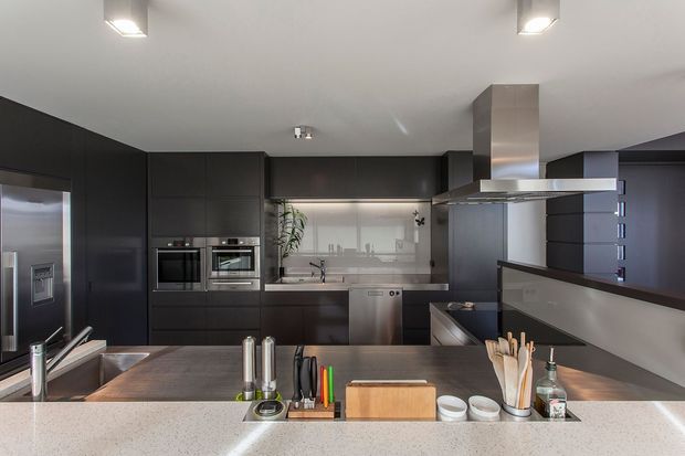 Contemporary kitchen viewed from the island, featuring dark matte cabinetry, integrated stainless-steel appliances, a pale stone island benchtop, stainless-steel rangehood, splashback glazing, and recessed and pendant-style ceiling lighting. The kitchen has a clean, sophisticated, high-contrast fit-out with a secondary dining or living area visible beyond the raised partition.