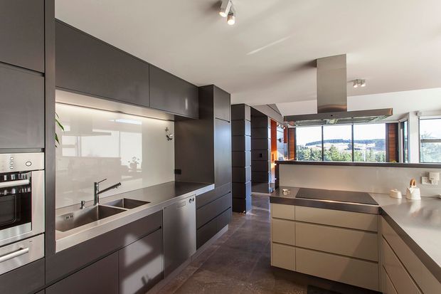 A contemporary galley-style kitchen with dark grey full-height cabinetry along the left wall, integrated appliances, a double-bowl sink, and a large central island with a projecting benchtop. The room has a light ceiling with track lighting, dark tiled flooring, stainless-steel appliances and rangehood, and broad glazing at the far end overlooking a leafy suburban landscape. A corridor or adjoining living area is visible beyond the kitchen.