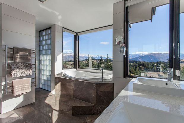 A bright, contemporary bathroom with a freestanding corner bathtub, separate built-in bath or vanity in the foreground, heated towel rail, glass-block partition, and large corner windows overlooking a mountainous landscape. The room features large dark stone-look floor and wall tiles, pale wall panels, and a sculptural tiled bath surround.