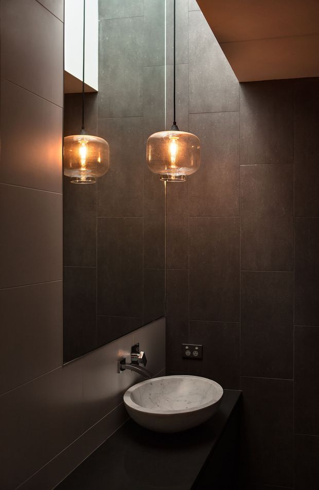 Moody, compact bathroom vanity with a round white stone vessel basin on a dark benchtop, wall-mounted chrome tapware, a large mirror, dark large-format tiled walls, and two warm-glowing pendant lights. The space has a dramatic, refined contemporary feel with strong contrast between the dark surfaces and illuminated amber glass pendants.