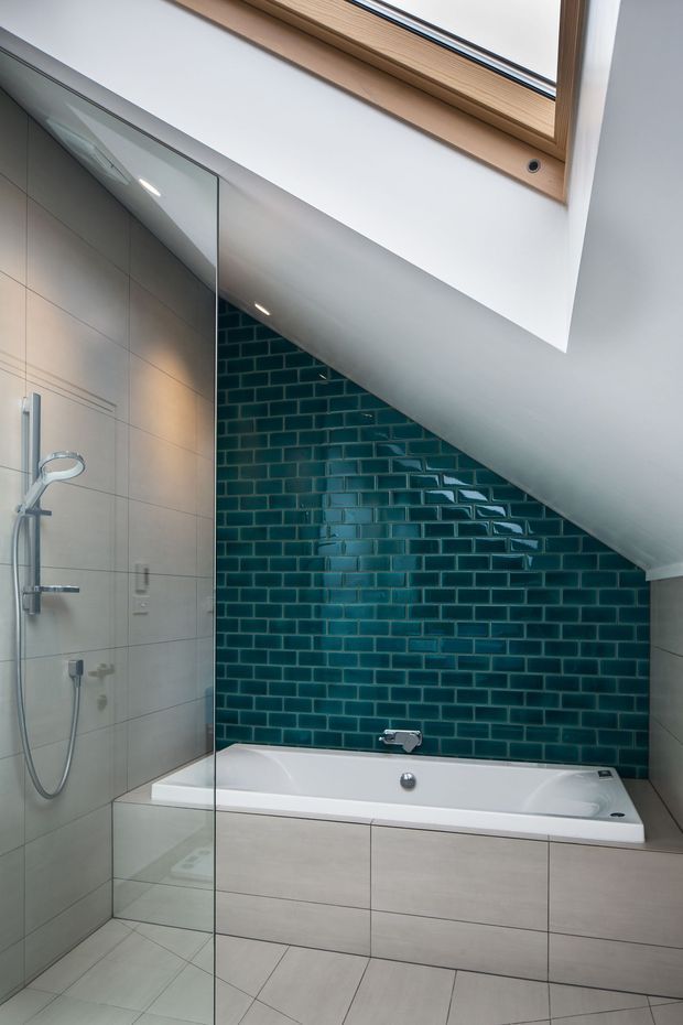 Compact contemporary bathroom beneath a sloping roof, featuring a built-in white bathtub, glass shower enclosure, pale large-format wall and floor tiles, and a striking glossy teal subway-tile feature wall. A timber-framed skylight brings natural light into the space.