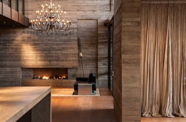 A dramatic, high-ceilinged contemporary living space featuring extensive warm timber wall cladding, a long linear fireplace, a sculptural black lounge chair, and a large ornate multi-arm chandelier. A substantial stone or concrete island/counter occupies the foreground, while full-height pleated drapery screens an opening on the right. The palette is warm brown, tan, black, and muted metallic, with strong textural contrast between timber, masonry, fabric, and polished flooring.