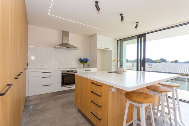 Bright contemporary kitchen with a timber-panelled wall of tall cabinetry, white flat-panel cabinets, a white herringbone-tile splashback, stainless-steel rangehood, concrete-look flooring and a large white island with timber cabinetry. The island includes casual bar seating, and full-height sliding glass doors open to an outdoor view.