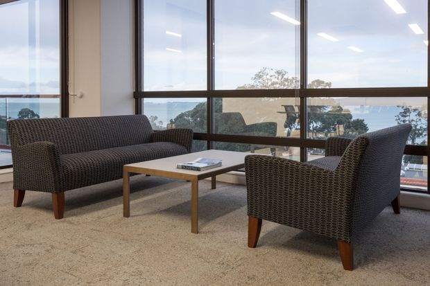 A bright, refined lounge or waiting area with a woven dark armchair and matching two-seat sofa arranged around a simple timber coffee table. The room has expansive floor-to-ceiling glazing with dark horizontal and vertical frames, carpet flooring, and views toward a wooded waterfront or landscape beyond. Linear ceiling lights and restrained furnishings create a calm, functional atmosphere.