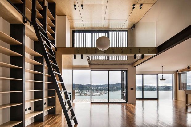 A spacious, double-height contemporary interior with polished timber flooring, extensive built-in open shelving and a leaning library ladder on the left. A mezzanine-level walkway with exposed dark structural beams overlooks the open living area. Floor-to-ceiling glazing provides expansive views across a body of water and surrounding hills. The ceiling combines light timber panels, exposed services and track lighting, with a large spherical pendant as a focal point.