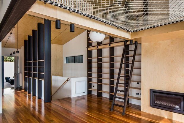 Interior view of a contemporary multi-level living space featuring extensive built-in dark timber shelving, a rolling library ladder, warm plywood wall and ceiling finishes, polished timber flooring, black structural columns, and a built-in fireplace. A rope mesh safety or play net spans the upper ceiling area, with a stair opening and glimpses of an adjoining room visible at left.