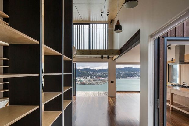 A tall, narrow contemporary interior with open built-in shelving in dark and light timber along the left wall, polished timber flooring, high ceilings, exposed structural beams, and pendant and track lighting. Full-height glazing at the far end frames a waterfront harbour and hilly urban landscape. A kitchen with timber cabinetry and a benchtop is visible through the opening on the right.