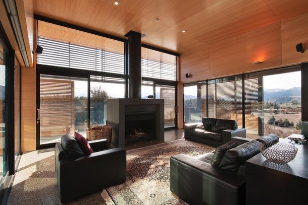 A contemporary living room with extensive floor-to-ceiling glazing, warm timber-lined walls and ceiling, dark structural columns, and horizontal timber blinds. Black leather sofas and armchairs surround a central built-in fireplace, with a large patterned rug and dark sideboard. The windows open to expansive mountain and suburban views, creating a bright, warm, indoor-outdoor atmosphere.