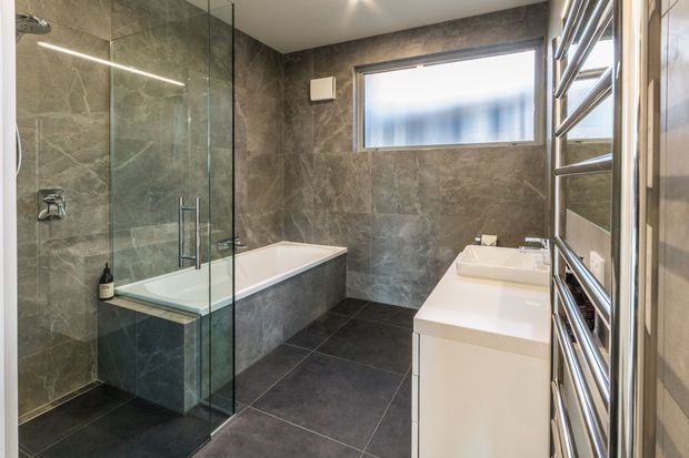 Contemporary bathroom with dark grey stone-look wall and floor tiles, a frameless glass walk-in shower, built-in bathtub, and a white floating vanity with integrated basin. A large chrome heated towel rail occupies the right side, while a frosted horizontal window provides natural light.