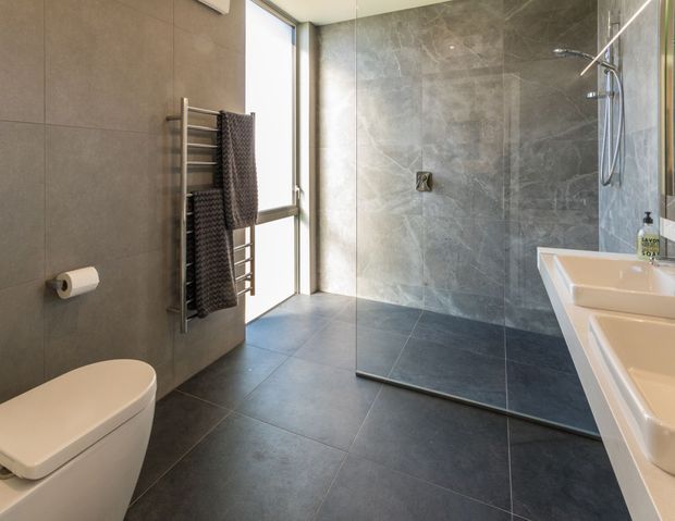 Contemporary bathroom with large-format dark grey stone-look wall and floor tiles, a frameless glass walk-in shower, white wall-mounted toilet, chrome heated towel rail, and a white double vanity. Tall glazing brings in natural light, creating a clean, refined atmosphere.