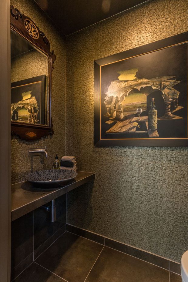 A compact, moody powder room with dark textured wallcovering, dark tiled flooring, and a floating dark vanity with a patterned vessel basin and wall-mounted chrome tap. An ornate dark-framed mirror reflects the basin, while a large framed artwork dominates the wall. Warm accent lighting creates an intimate, dramatic atmosphere.