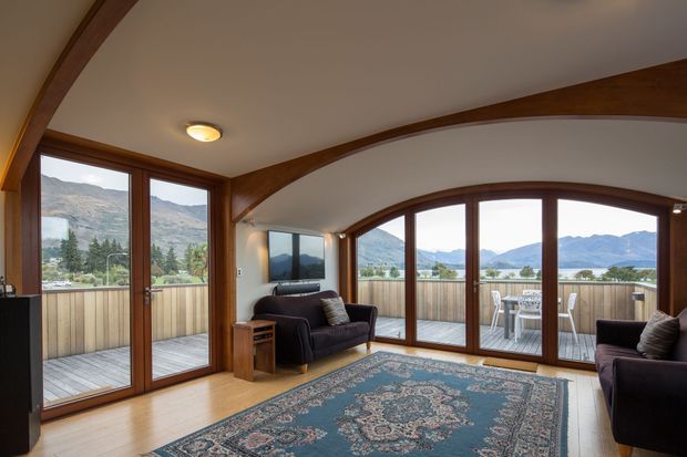 A warm living room with expansive timber-framed glazing and curved exposed timber beams, opening onto a balcony with mountain and lake views. The room contains dark upholstered sofas, a patterned area rug, timber flooring, a wall-mounted television, and a small side table. The space feels like a refined mountain retreat with strong indoor-outdoor connection.