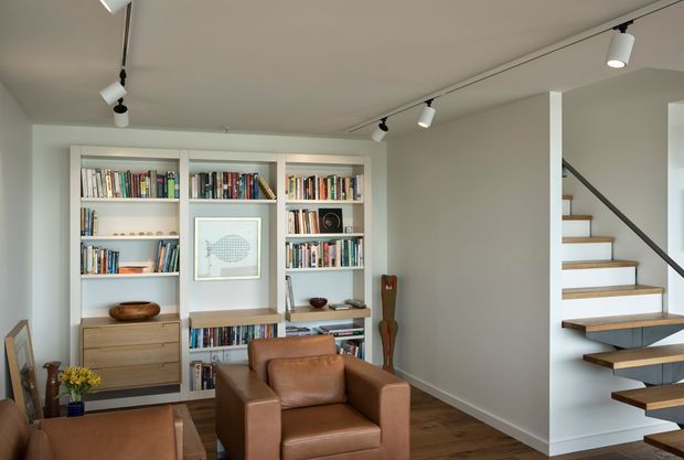 A bright, compact living room or reading area with a built-in white bookcase filled with books, a tan leather armchair, timber flooring, and a framed artwork. Open floating timber stairs with a dark metal handrail rise along the right side. Track lighting runs across the smooth white ceiling, creating a clean, contemporary interior.
