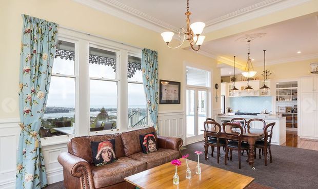 A bright, traditionally styled open-plan living and dining area with a kitchen visible beyond. The room features bay windows with decorative patterned valances and floral curtains, pale yellow walls, white wainscoting and ornate cornices, a brown leather sofa, dark timber dining furniture, and a wood coffee table. Large windows overlook a waterfront or harbour setting, while classic chandeliers and pendant lights add decorative character.