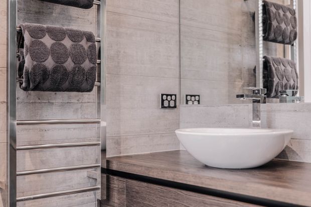 Close-up view of a contemporary bathroom vanity with a white vessel basin, wall-mounted chrome tapware, dark timber-look vanity cabinetry and benchtop, pale large-format concrete-look wall tiles, and a stainless-steel heated towel rail. A mirrored cabinet reflects the textured towel and part of the vanity.