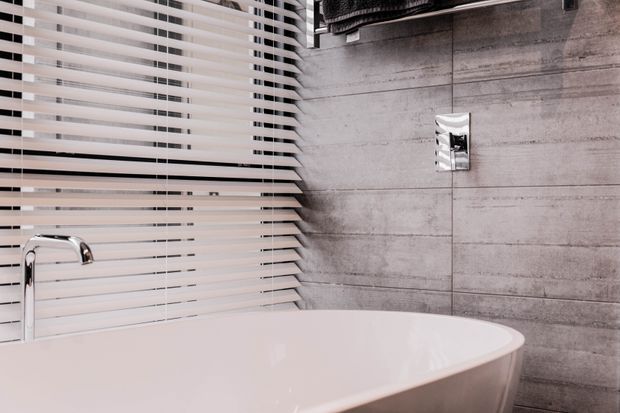 Close-up of a contemporary bathroom featuring a freestanding white bathtub in the foreground, a chrome wall-mounted tap, grey concrete-look wall tiles, and horizontal white blinds covering the window. The space has a restrained, monochromatic palette with polished modern fixtures.