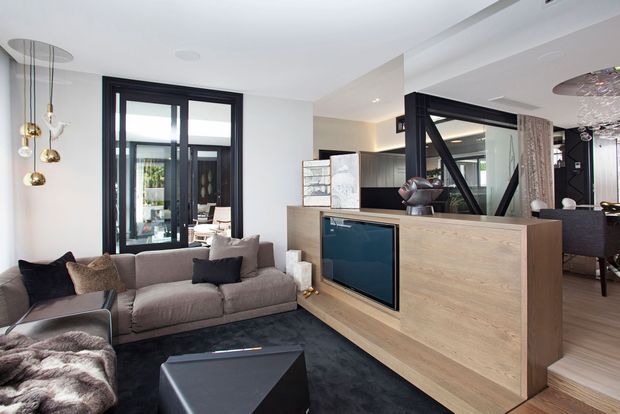 Contemporary open-plan living room with a large grey sectional sofa, dark rug and geometric coffee table. A substantial light timber built-in entertainment and storage unit divides the living area from the dining space, with a recessed television and decorative objects. Black-framed glazed doors and partitions, white walls, contemporary pendant lighting and glimpses of the dining area complete the refined interior.