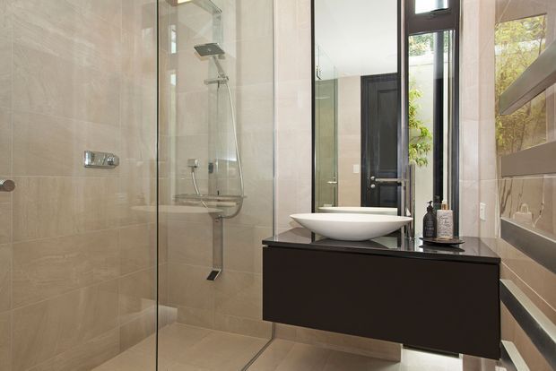 Contemporary bathroom featuring large beige stone-look wall and floor tiles, a frameless glass shower enclosure with chrome fixtures, a white vessel basin on a dark floating vanity, and a tall black-framed window or glazed door. A large mirror reflects the vanity and shower area.