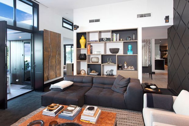 Bright contemporary living room with a large dark sectional sofa, white armchair, patterned rug, and a substantial timber coffee table. A built-in open shelving unit displays books and decorative objects, while full-height glazing and a dark entry corridor create a strong contrast. The room opens toward a dining or kitchen area in the background and includes a dark textured feature wall.