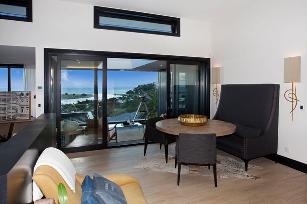 A refined contemporary living room with pale timber flooring, white walls, black-framed floor-to-ceiling sliding glazing, and clerestory windows. The room contains a round timber dining or sitting table with dark upholstered chairs, a tall charcoal lounge chair, and a partially visible mustard armchair. Through the glazing, a coastal landscape and pool terrace are visible. Black cabinetry or joinery is visible at left, and sculptural wall lighting adds a decorative accent.
