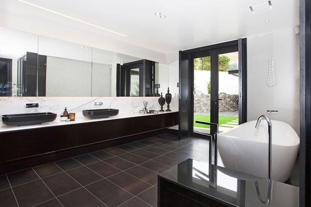 A spacious contemporary bathroom with twin vessel basins on a dark floating vanity, a large wall mirror, a freestanding white bathtub, dark tiled flooring, and black-framed glazed doors opening to a landscaped courtyard. The palette is monochromatic with white surfaces, charcoal joinery, and polished dark finishes.
