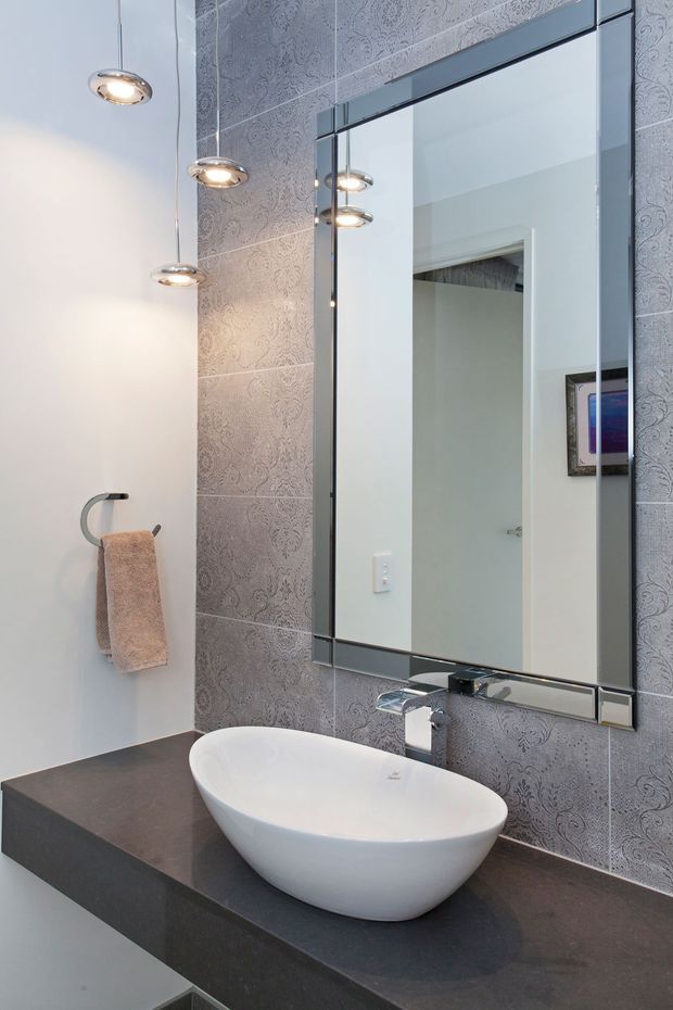 Contemporary bathroom vanity featuring a sculptural white vessel basin on a dark stone benchtop, a large framed mirror, patterned grey wall tiles, chrome tapware, and pendant lights.