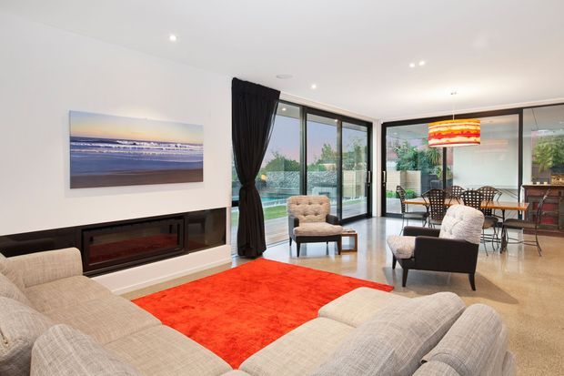 Open-plan contemporary living and dining area with a large cream sectional sofa, bright red area rug, built-in dark media and fireplace joinery, and floor-to-ceiling glazed doors opening to an outdoor pool area. A dining table sits near the glazing, with a colourful pendant light and beach artwork adding visual accents.