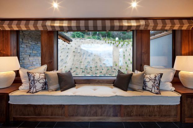 A cozy built-in window seat beneath a wide striped Roman blind, framed by dark timber panelling and cabinetry. The seat is upholstered in cream fabric with layered neutral cushions, flanked by table lamps. A stone retaining wall and planted slope are visible through the central window.