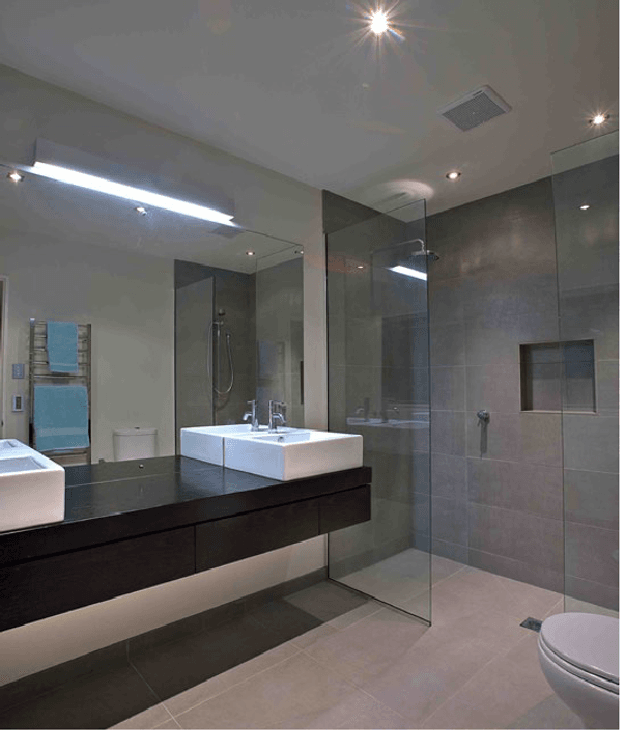 A contemporary bathroom with large-format grey tiled walls and floor, a frameless glass walk-in shower, floating dark timber vanity with white rectangular basins, mirrored wall panels, recessed shower niche, and concealed ambient lighting. A toilet is visible at the right.