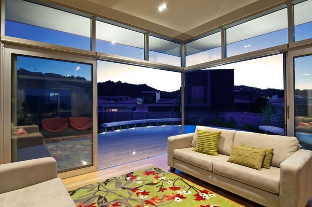 A contemporary living room with expansive floor-to-ceiling glazing and sliding glass doors opening onto a deck or terrace. The room contains a pale upholstered sofa and armchair, colourful patterned rug, and bright accent cushions. Evening light reveals a suburban hillside setting beyond the glazed walls, while recessed ceiling lighting keeps the interior softly illuminated.