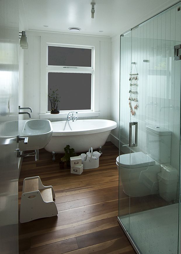 Bright contemporary bathroom with a freestanding oval bathtub beneath two windows, a wall-mounted basin, and a large frameless glass shower enclosure. The room features white panelled walls, white sanitaryware, dark timber floorboards, and brushed metal fixtures, with a small amount of decorative greenery and storage.