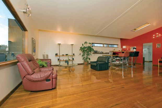 Open-plan contemporary interior with polished timber flooring, a spacious living area, large corner glazing, leather recliner seating, glass shelving and coffee table, and a compact kitchen or bar zone at the rear. A vivid red feature wall adds strong contrast to the otherwise light neutral room.