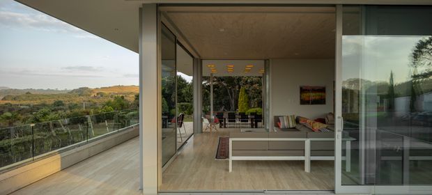 A contemporary indoor-outdoor living space viewed from a broad terrace, with floor-to-ceiling sliding glass doors, pale timber flooring, a white built-in or low-profile seating area, and a covered ceiling. The glazing opens toward a landscaped valley view, while the interior includes a compact seating zone and a visible dining or lounge area beyond.