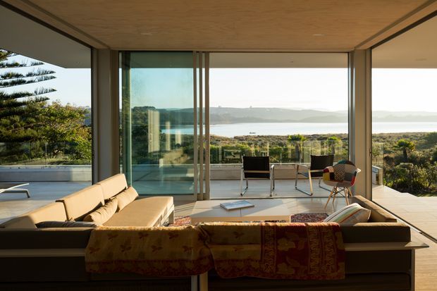 A modern living room opening onto a spacious terrace, with full-height sliding glass walls framing a broad coastal water view. The room features a low-profile sectional sofa, timber ceiling, pale flooring, minimalist lounge chairs, and a pool or reflecting surface visible to the left. The interior is strongly connected to the outdoor landscape through extensive glazing.