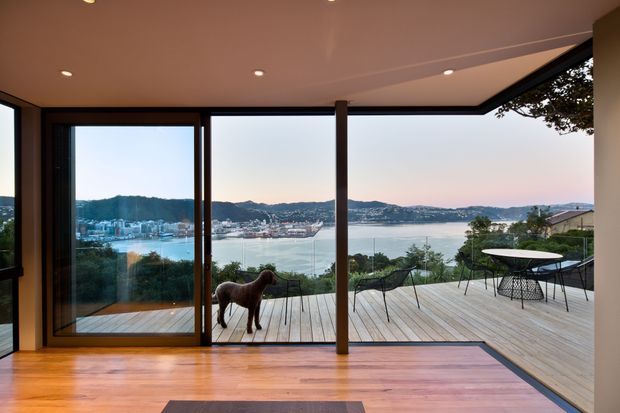 Contemporary indoor-outdoor living space opening onto a generous timber deck, with full-height glazing framing a broad harbour and hillside view. The interior has warm timber flooring, a smooth pale ceiling with recessed downlights, dark slender window and door frames, and a simple dining setting on the deck. A sculptural dog is visible indoors, while the main architectural focus is the seamless connection between the living area and elevated waterfront outlook.
