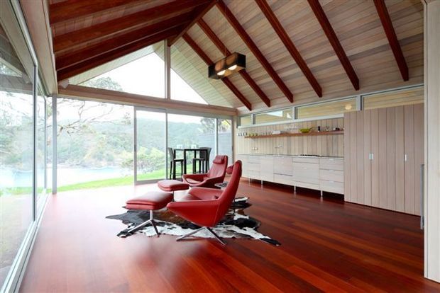 A bright, open-plan interior lounge and dining area with floor-to-ceiling glazing overlooking a leafy landscape. The room features a dramatic pitched ceiling with exposed dark timber rafters, pale timber ceiling boards, extensive built-in timber cabinetry and shelving, and a rich dark red-brown timber floor. Two red leather lounge chairs with matching ottomans sit on a cowhide rug, with a compact dining table near the glazing.