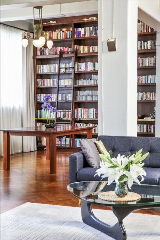 A refined living room and library area with extensive dark timber built-in bookshelves, a polished timber desk, hardwood flooring, a deep blue upholstered sofa, and a sculptural glass coffee table. Pendant lights hang near sheer curtains, while flowers add a soft decorative accent. The space has a warm, book-lined, sophisticated atmosphere.