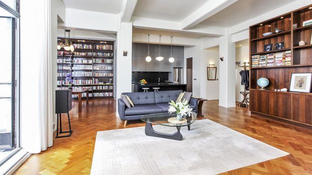 Bright open-plan living room with a dark blue sofa, sculptural coffee table, large pale rug, and extensive dark timber built-in shelving displaying books and objects. The space opens to a compact black kitchen and a library or study area, with polished herringbone timber flooring, white walls, exposed structural beams, and large industrial-style steel-framed windows.