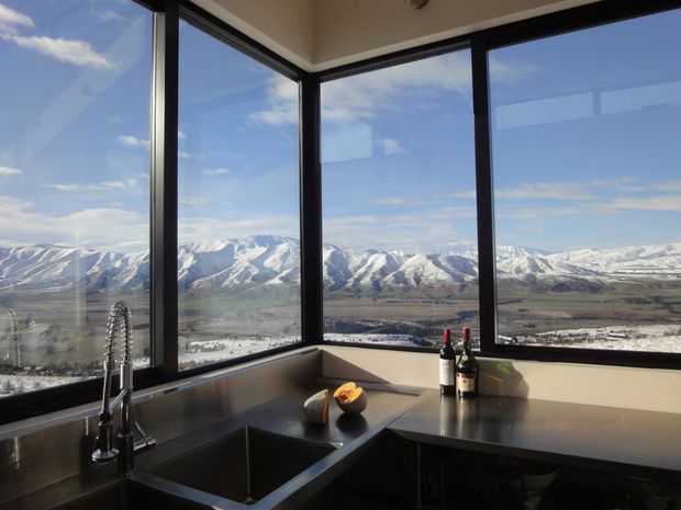 A contemporary kitchen corner with dark cabinetry and a dark stone benchtop, positioned beside expansive black-framed corner windows. A deep undermount sink with a tall spring-style tap occupies the left side, while wine bottles and fruit sit on the benchtop. The glazing provides a dramatic panoramic view of snow-covered mountains and a valley beyond.