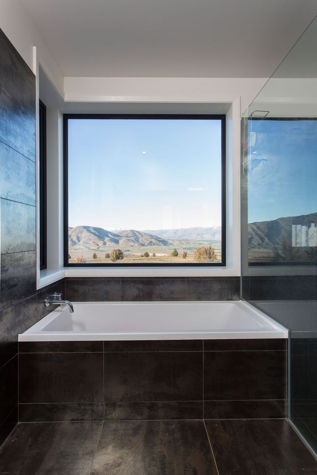 Contemporary bathroom centred on a built-in white bathtub beneath a large black-framed picture window. Dark glossy stone- or porcelain-look wall and floor tiles create a dramatic finish, while a full-height mirror or glass panel reflects the mountain landscape outside. The expansive glazing looks onto a dry rural valley with layered hills.
