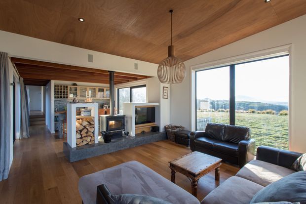 A bright, open-plan living room with warm timber flooring and a timber-lined ceiling. The room features a central double-sided fireplace with exposed firewood storage, a television niche, leather and upholstered sofas, a rustic wooden coffee table, and a large pendant light. Full-height glazing provides expansive views of the surrounding landscape. A kitchen or dining area is visible beyond the fireplace.