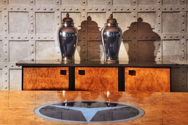 A dramatic, richly detailed interior vignette featuring a long dark-framed sideboard with glowing amber burl-wood cabinet fronts, topped by two polished silver urns. The backdrop is a rugged metallic or stone-look panelled wall with prominent rivets, while a highly polished warm timber table with a circular inset dominates the foreground. Strong directional lighting creates pronounced shadows and an opulent, industrial-inspired atmosphere.