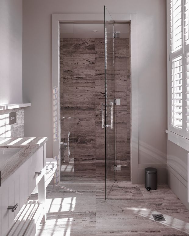 Bright, narrow contemporary bathroom viewed toward a glass-enclosed walk-in shower. The shower and floor are clad in large-format brown-grey marble-look tiles, with a frameless glass door and chrome hardware. A white vanity with a stone-look benchtop is partially visible on the left, while white plantation shutters on the right admit strong sunlight. A compact dark bin and floor drain complete the scene.