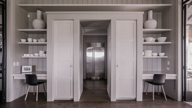 A refined built-in wall with symmetrical open shelving, display ceramics, framed artwork, and two compact desk workstations. Central double doors open to a secondary space containing a stainless-steel refrigerator. The room features vertical grey wall panelling, white painted cabinetry and trim, dark timber flooring, and black chairs, creating a calm, tailored interior.