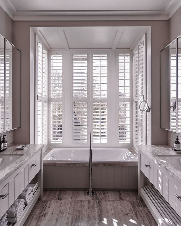 Bright, symmetrical bathroom with a freestanding bathtub positioned beneath a wall of white plantation shutters. Matching white vanity units with marble-look benchtops flank the room, while large mirrors reflect the cabinetry and shutters. The space has a grey and white palette, panelled walls and ceiling, and timber-look flooring.
