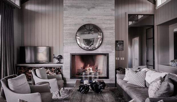 A sophisticated living room centred around a large contemporary fireplace with a grey textured feature wall and round mirror above. Vertical dark wall panelling, tall curtains, built-in firewood storage, a television console, and black-framed internal glazing frame the space. Two armchairs and a large grey sofa surround a low coffee table, creating a refined, cosy lounge with a neutral grey palette and warm firelight.
